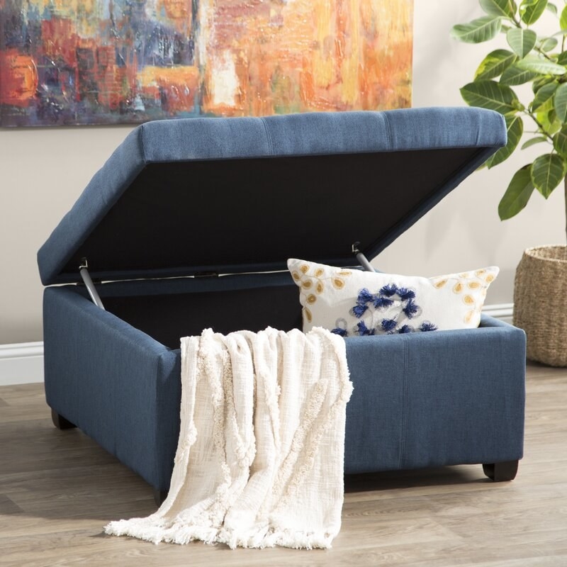 A dark blue, square ottoman with the top open displaying a pillow and blanket inside its storage feature