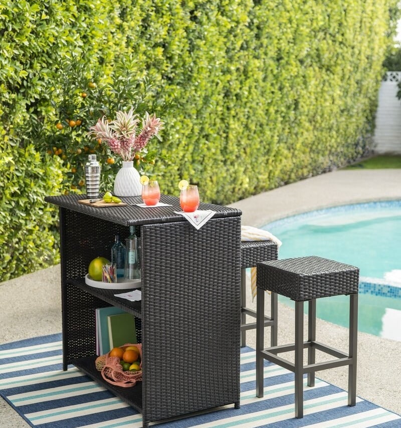 A wicker outdoor bar with two shelves and two stools displayed next to a pool