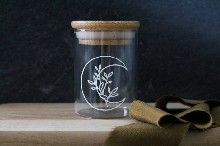 a small clear glass jar with a crescent moon animation on the front with flower sitting inside the crescent and a bamboo top