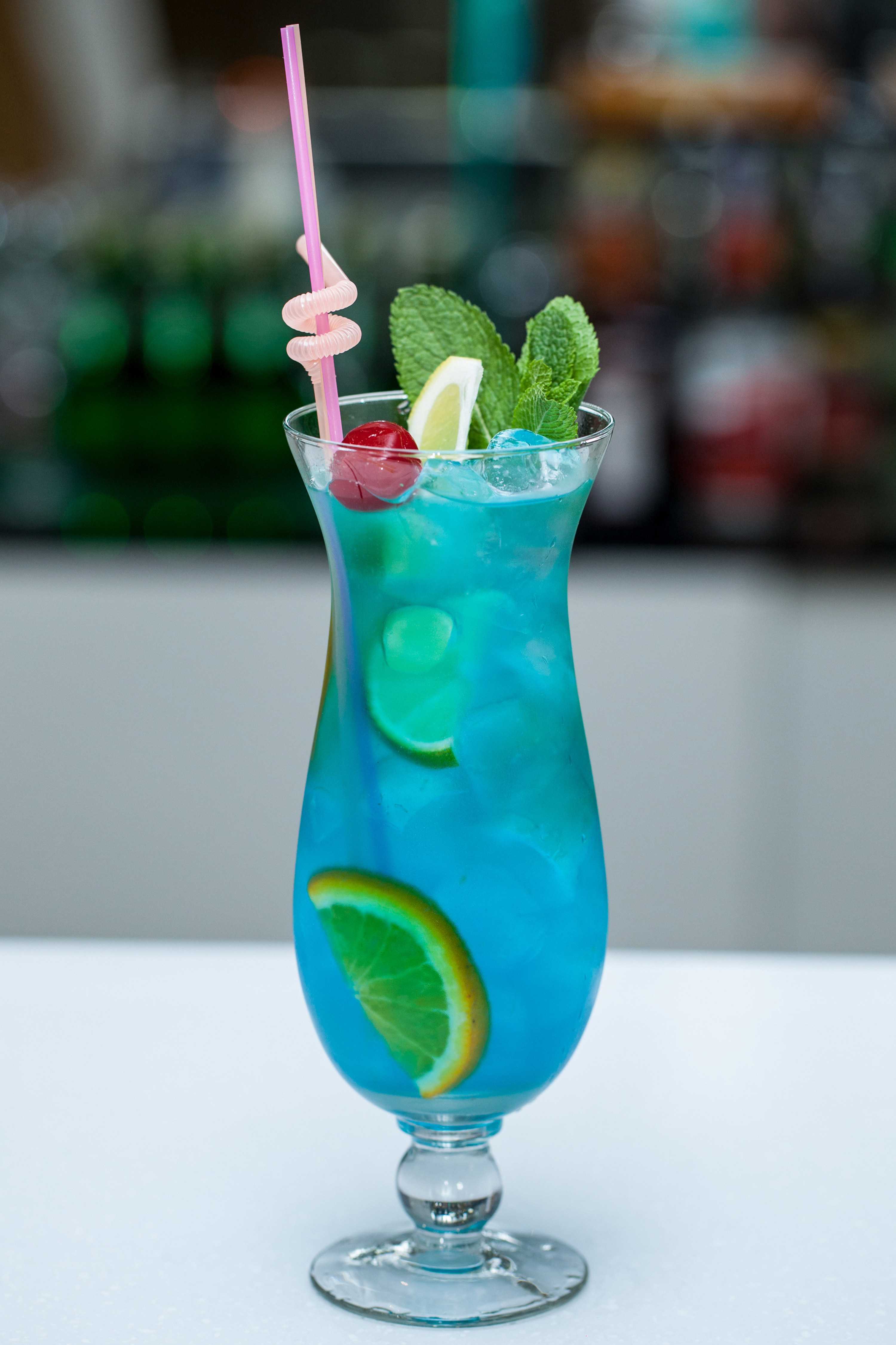 Glass filled with blue cocktail, a slice of lemon, mint leaves, a cherry, and a pink straw.