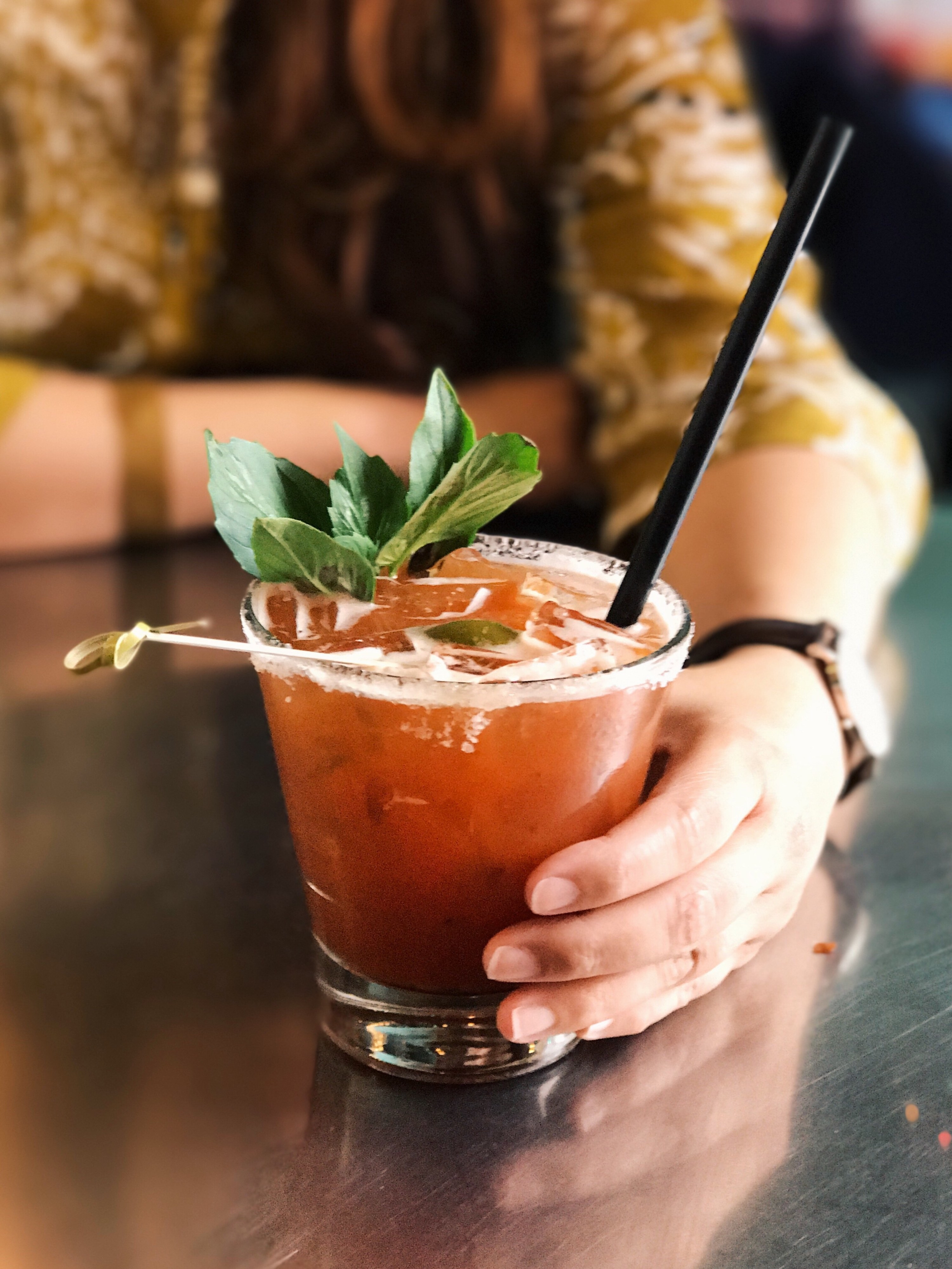 Person holding a Bloody Mary topped with herbs and a dark straw