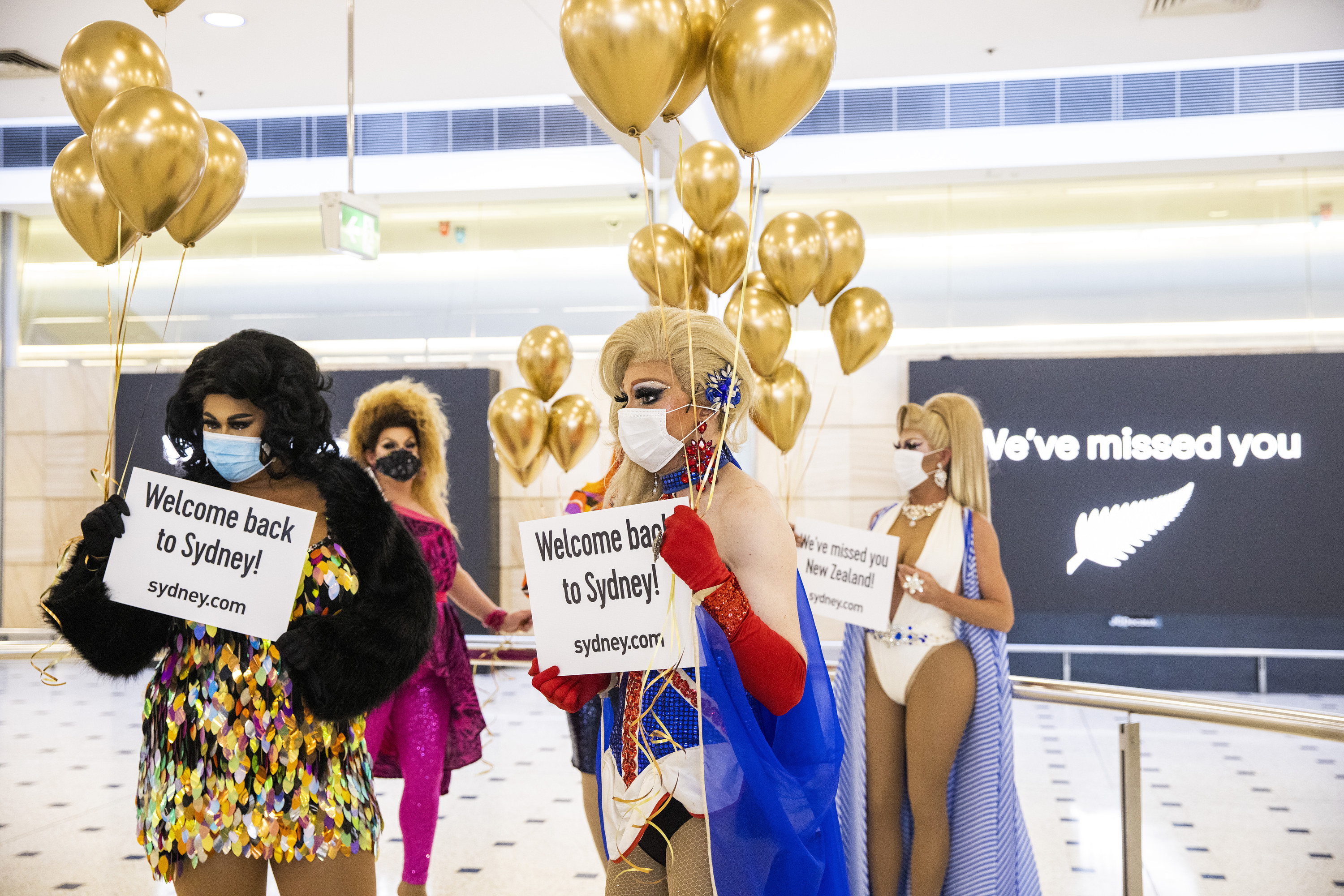 Drag queens holding signs that say &quot;welcome back to Sydney!&quot; and balloons