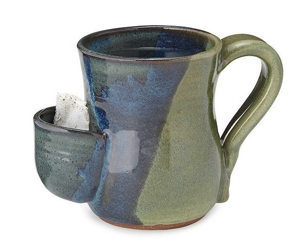 a green and blue stoneware mug with a curved handle and a front compartment with a tea bga inside 