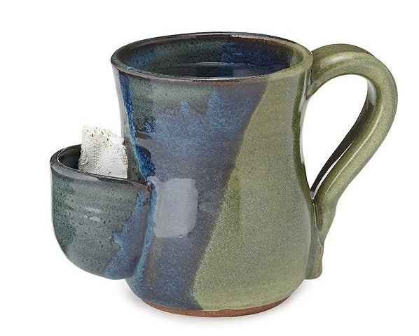 a green and blue stoneware mug with a curved handle and a front compartment with a tea bga inside 