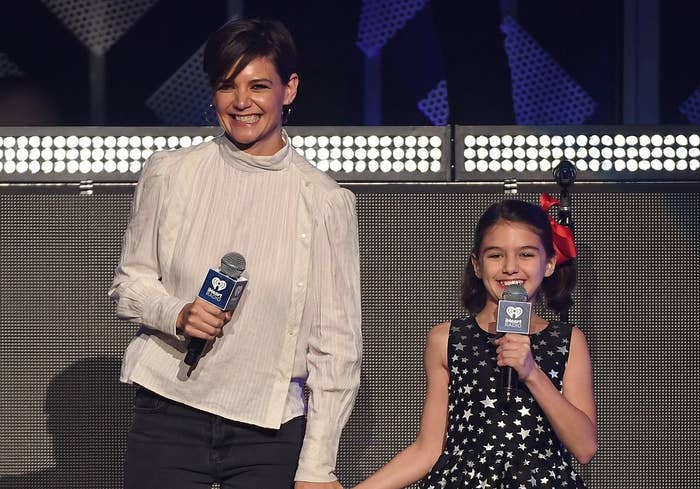 Katie and Suri hold hands while speaking onstage at an event