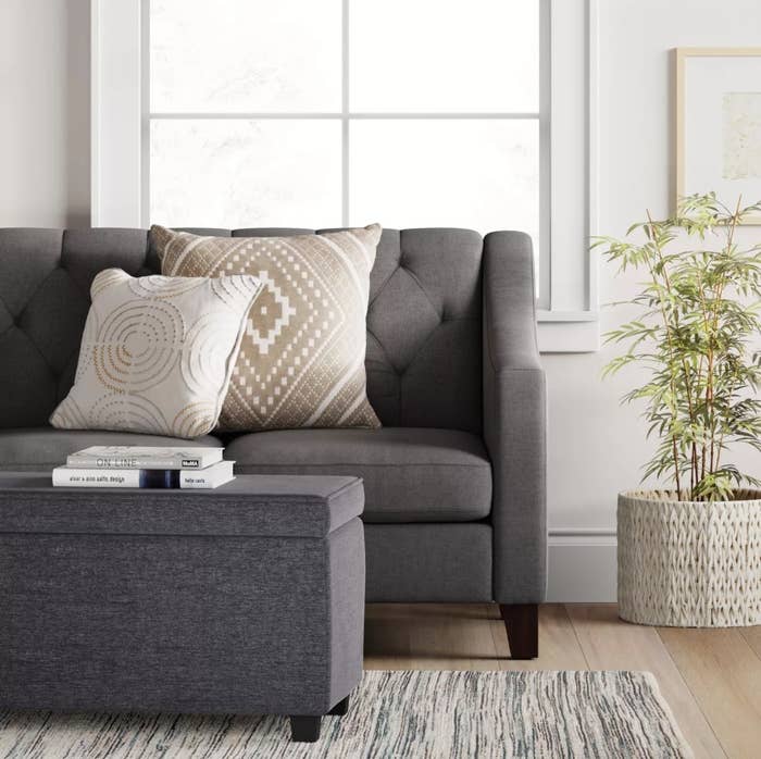 The gray storage ottoman in front of a pleated couch
