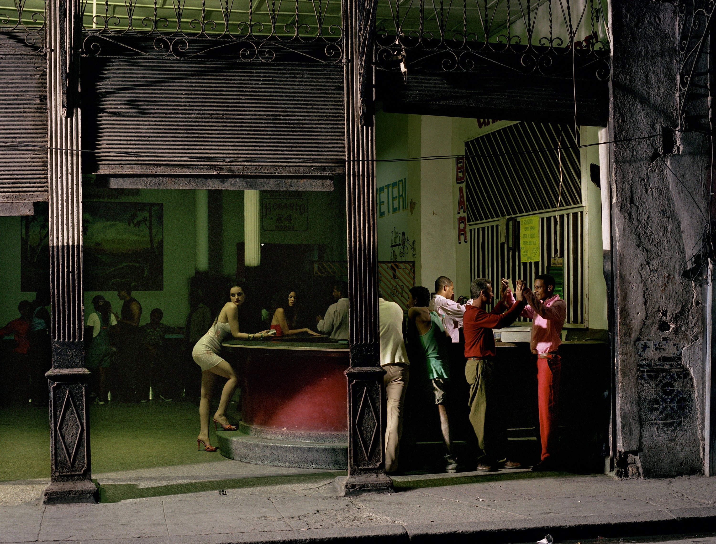 A young woman standing alone at a bar looks out past the camera while a group of men stand together to the right