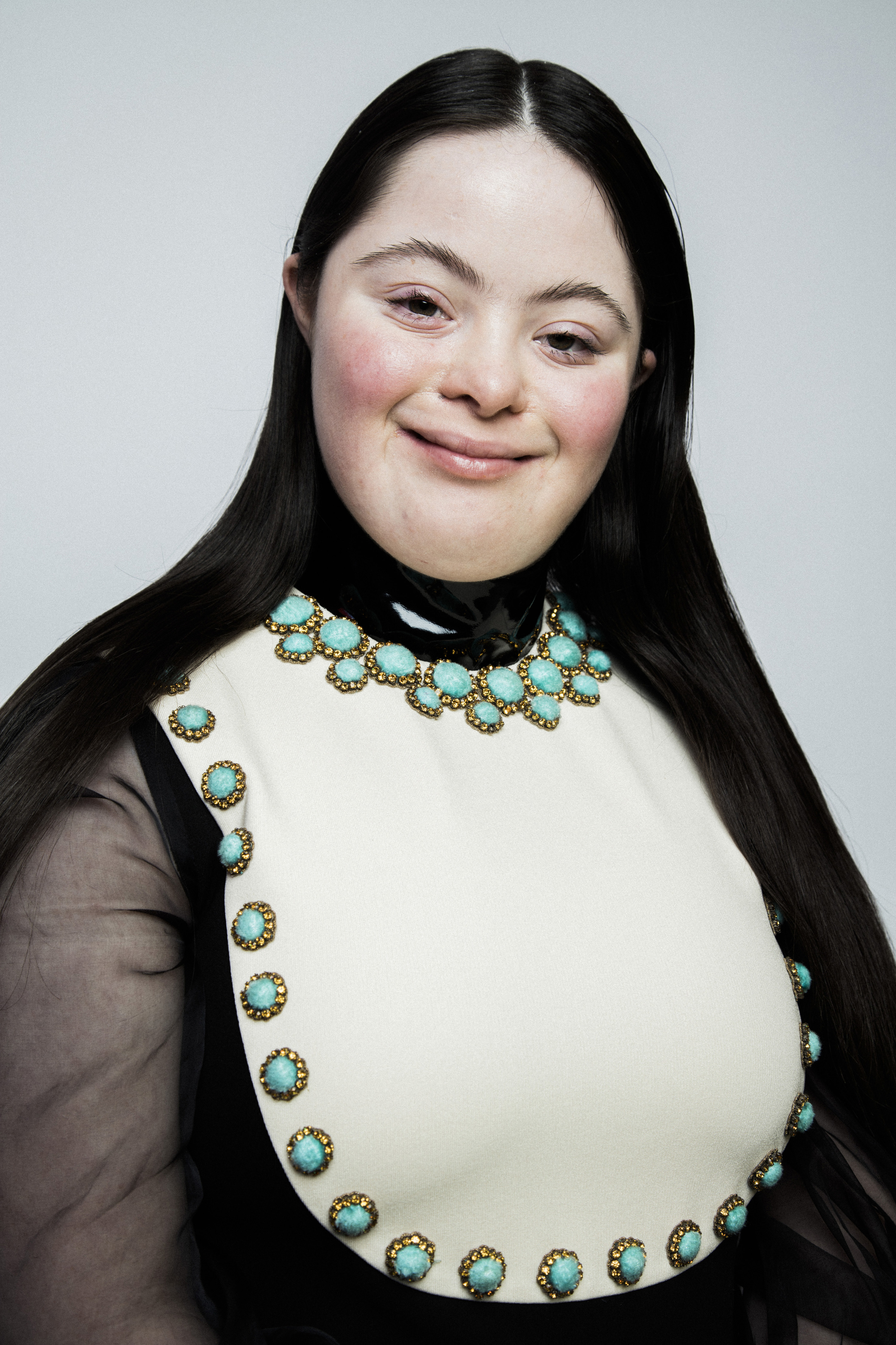 A young woman with long hair wears a studded bib and looks at the camera