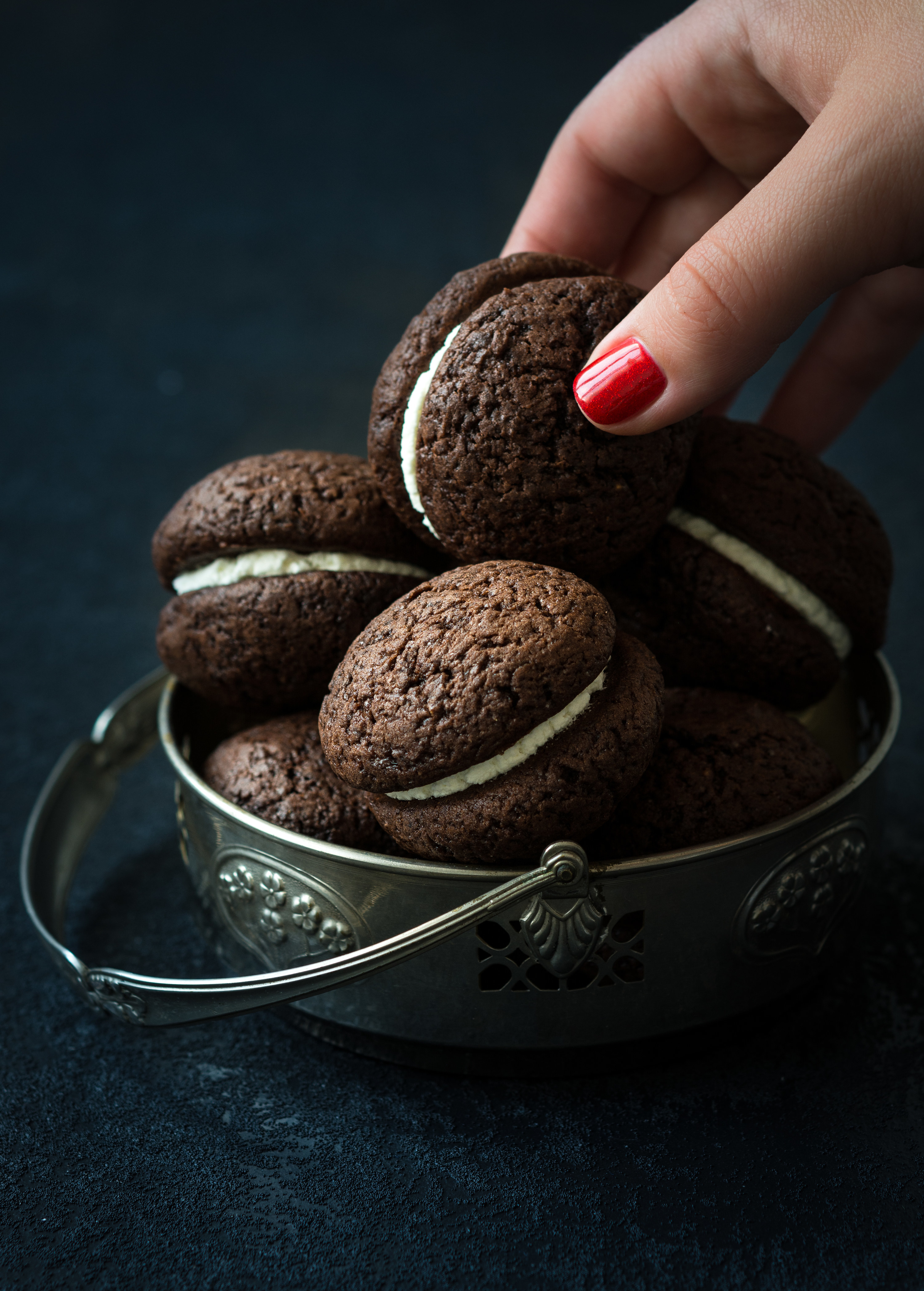 Manicured hand grabbing a dark whoopie pie consisting of chocolate cookies and a cream filling; there is also a metal container containing four more whoopie pies