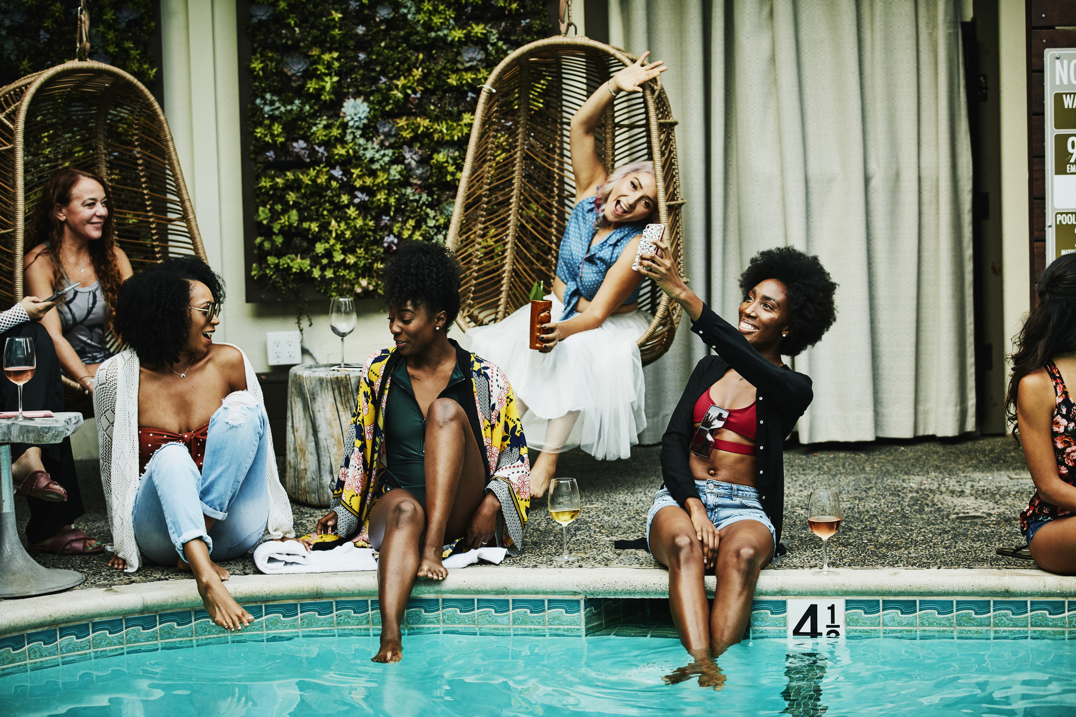 Five  female friends hang out poolside, smiling, taking photos, and sitting next to glasses of wine