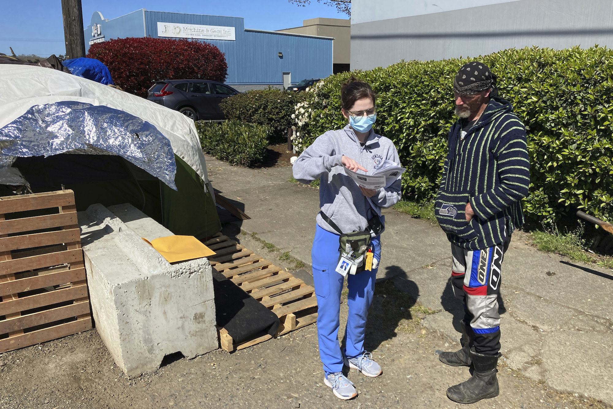 A woman in blue medical scrubs points to a piece of paper with COVID vaccine information while a man who lives at an encampment for people who are homeless looks on.