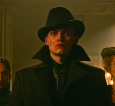 Kaz wears a hat and coat and stares straight ahead