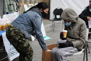 灰色连帽衫的一名妇女坐在金属椅子户外并且在获得一个covid疫苗以后在洛杉矶角度遮住的无家可归的人民的灰姑娘。