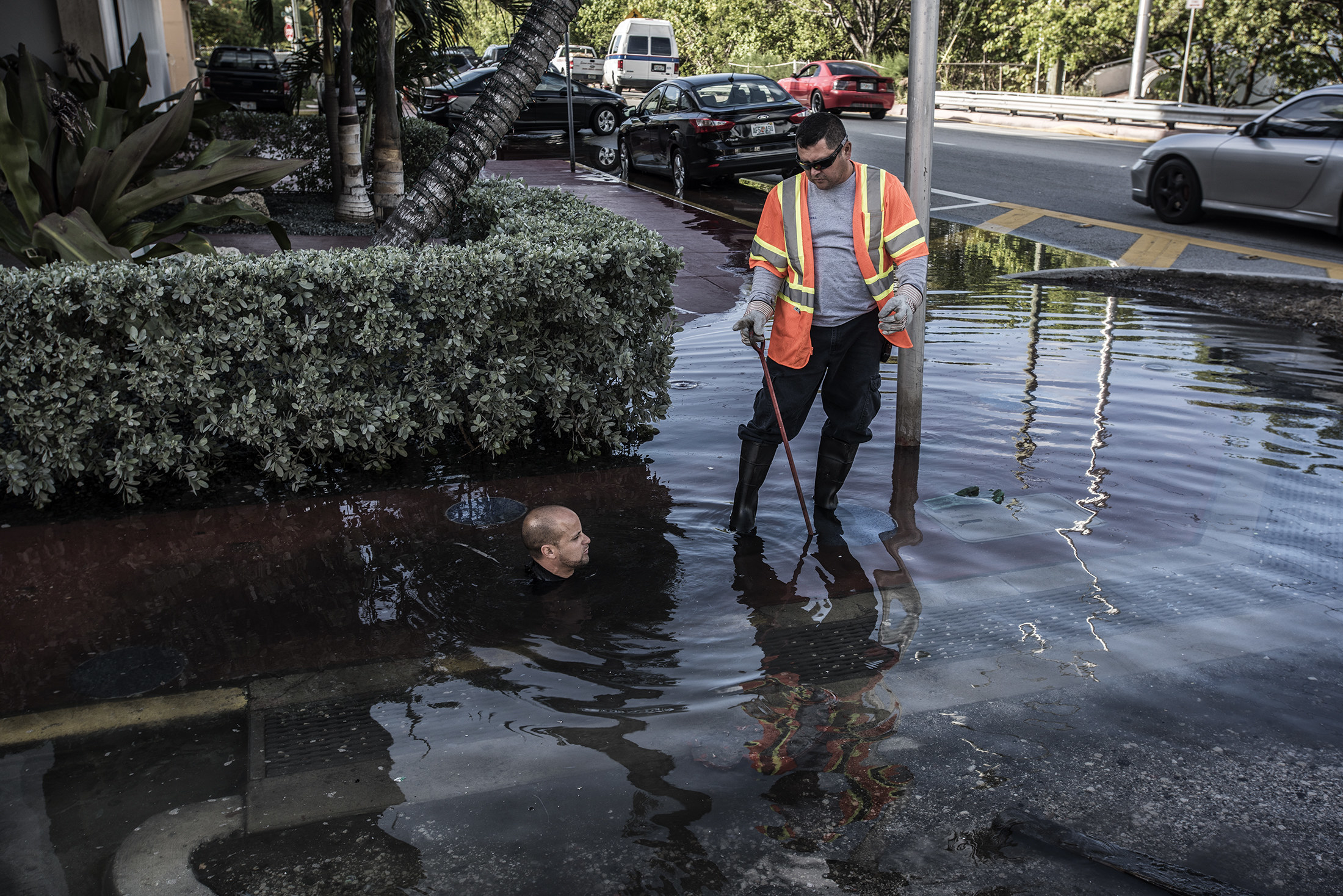 A worker mostly submerged in water with only his head showing on the street, next to another worker standing next to him with water up to his ankles 