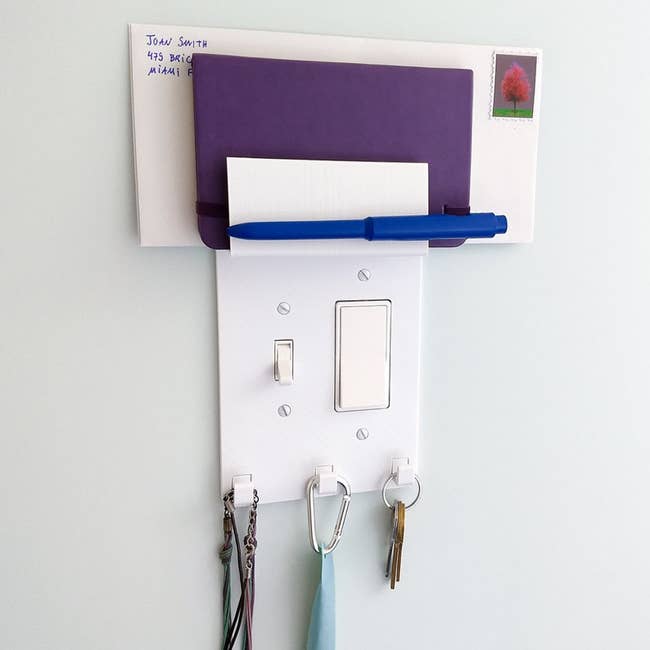 A white plastic cover for the lightswitch with three hooks for keys and a small shelf at the top for mail and a tiny notebook 