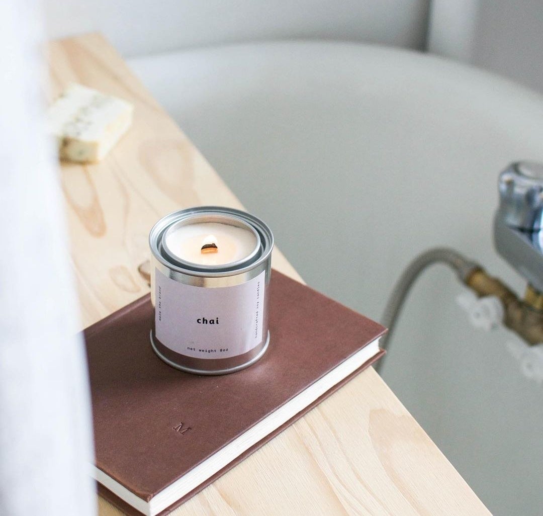 A lit candle in a metal tin resting on a book near a bathtub