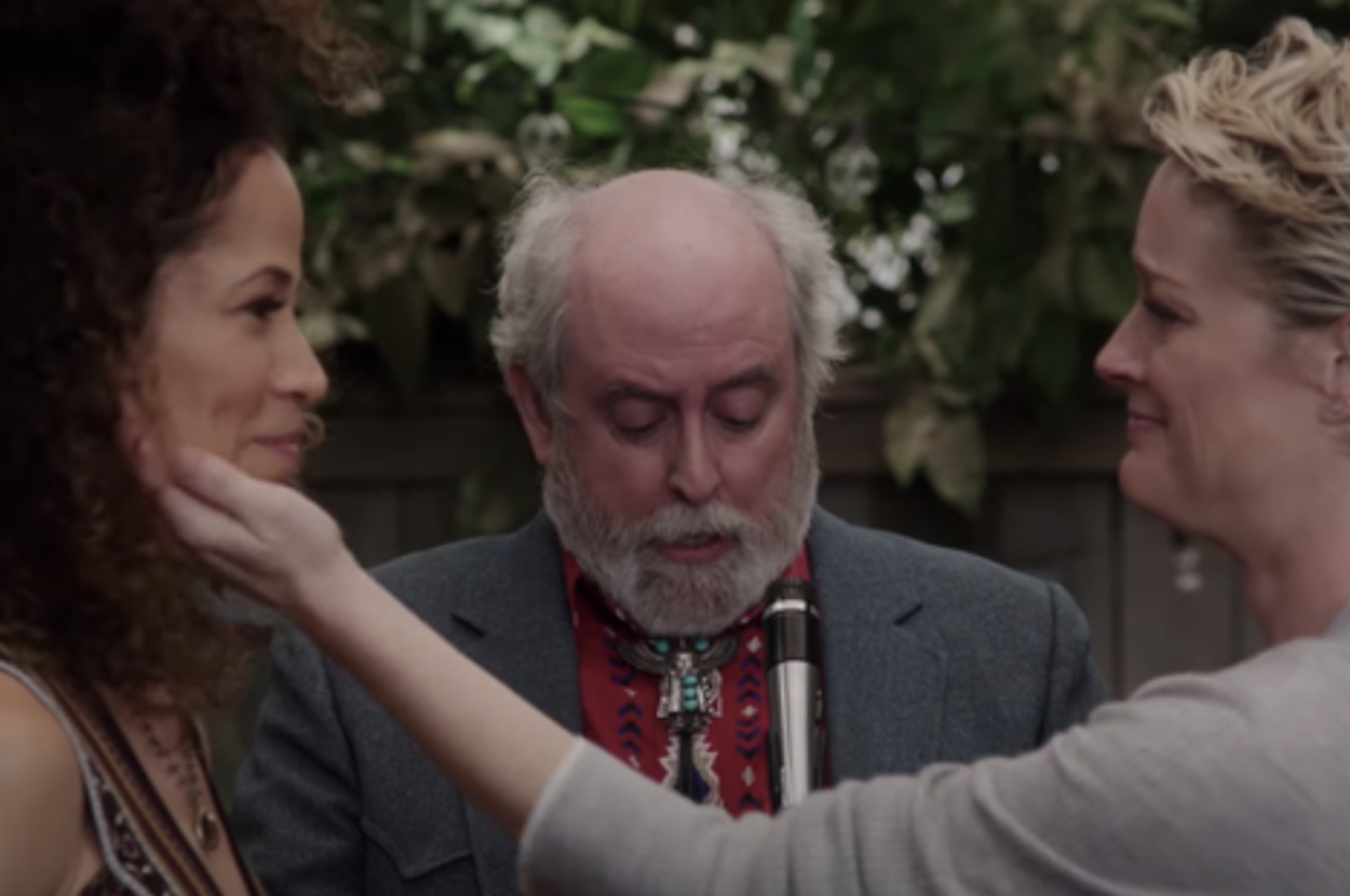 Two people are engaged in a tender moment outdoors, with a bearded person in the background, holding a microphone, possibly officiating