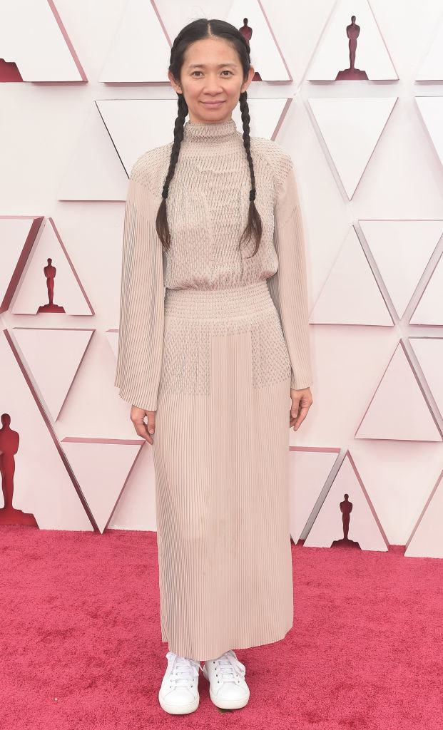 Chloé Zhao in pigtails and wearing a beige long-sleeved, high-neck gown with white sneakers