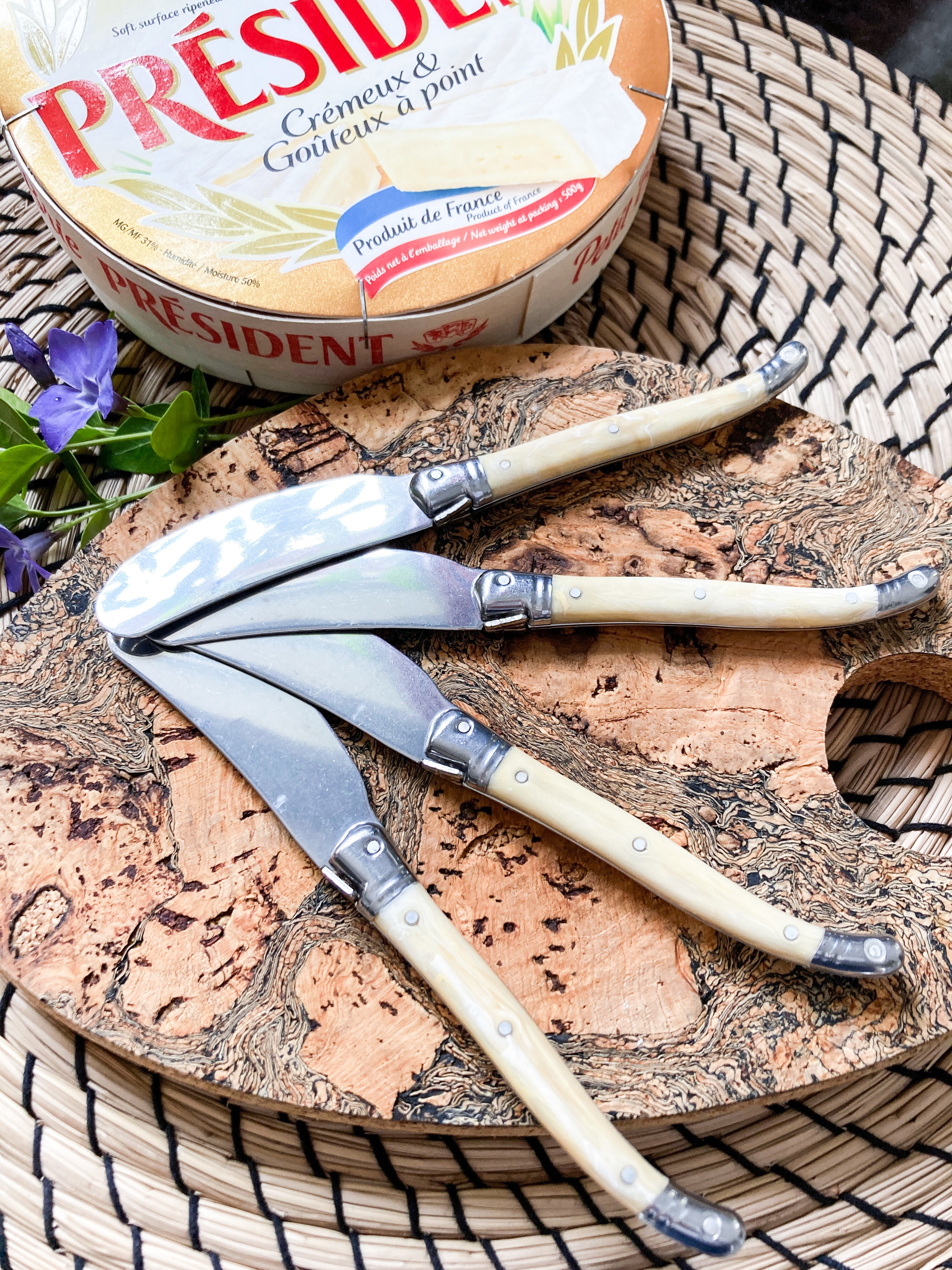 A flatlay of four charcuterie knives on a cork trivet next to creamy brie