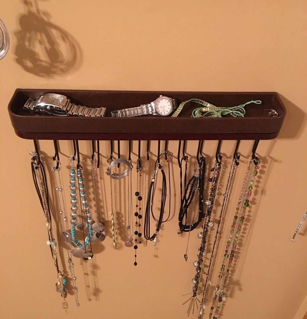 a reviewer photo of a wall-mounted jewelry organizer with a top tray and multiple hooks with necklaces hanging on them 