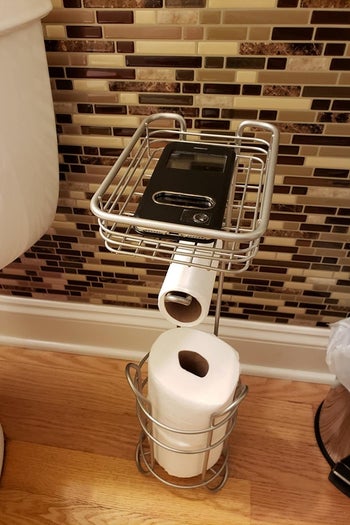 a reviewer photo of the wire stand in silver with a cell phone sitting in the top tray and two rolls of toilet paper sitting in the bottom section 