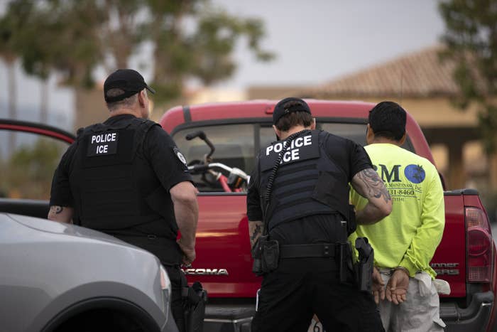 Two officers wearing vests with &quot;police ICE&quot; patches stand by a man whose hands are cuffed behind his back