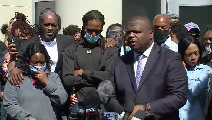 A group of Black people, frowning, grieving, and their arms crossed, stand around news station microphones