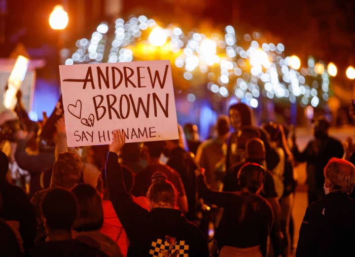 A demonstrator in a crowd holds up a sign that says "Andrew Brown, say his name" and has small drawings of a heart and a broken heart