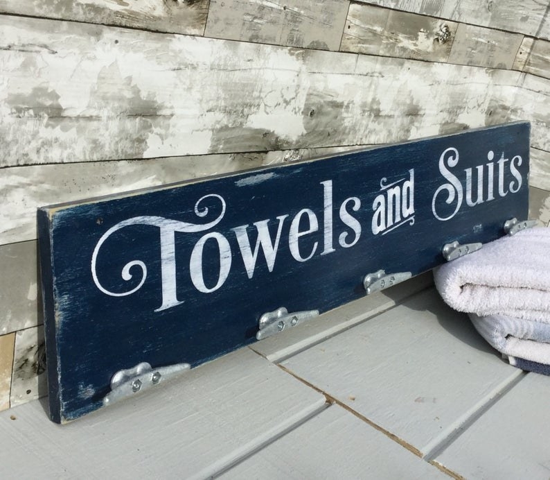 Rectangular wooden sign with metal hooks. The sign says &quot;Towels and Suits&quot; in a nautical script. 