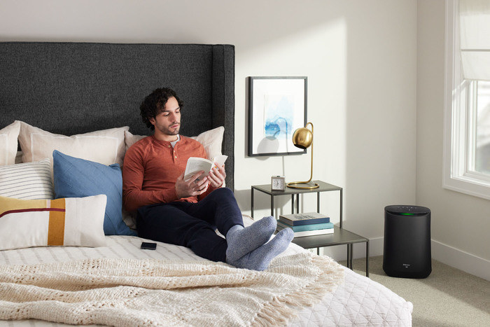 Model sitting on bed using smartphone while white air purifier sits in corner