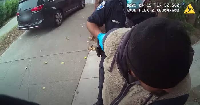 A man in a beanie is seen on police body camera footage being held by the arm of an officer wearing protective gloves and a mask