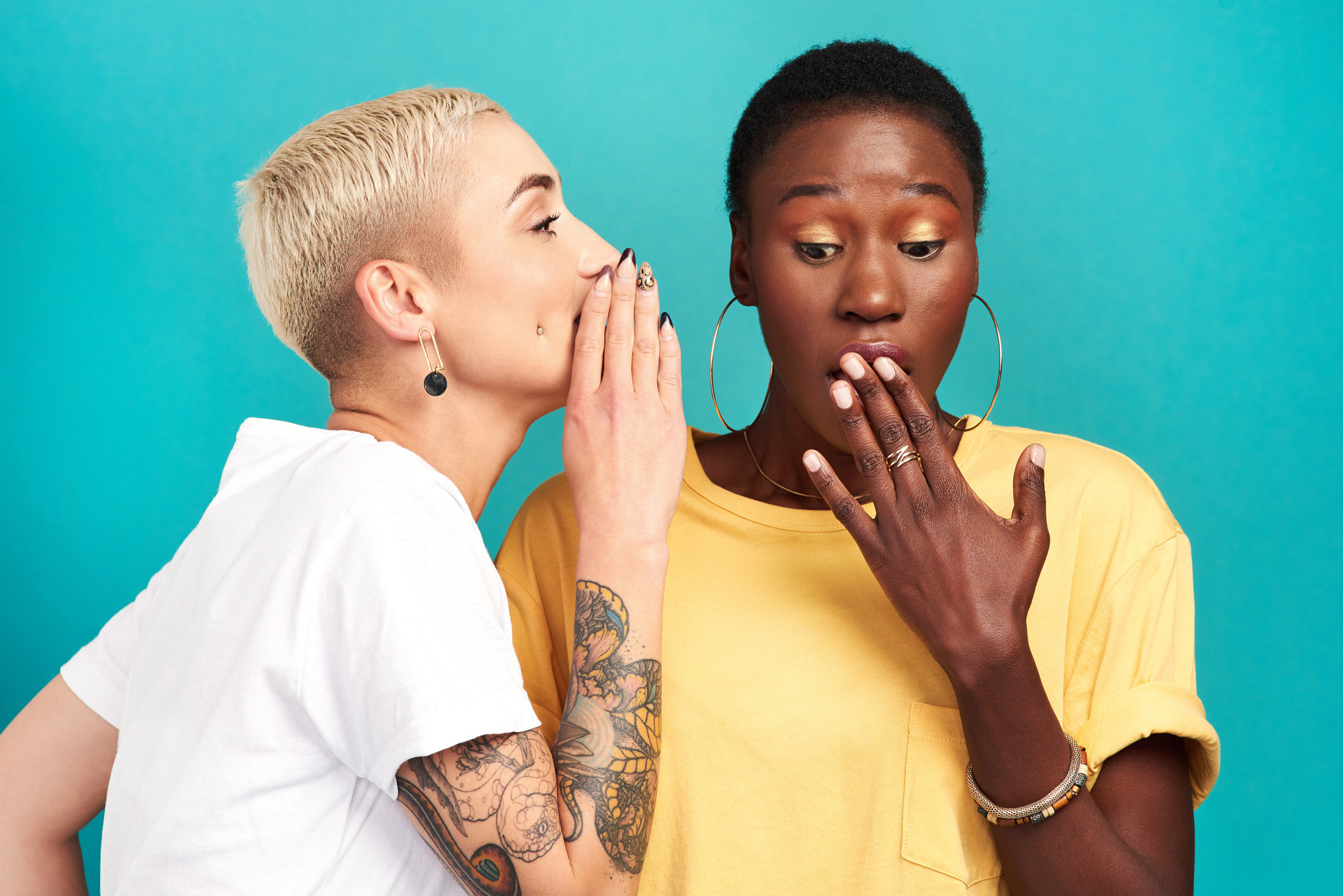 Studio shot of a young woman whispering in her friendâ€™s ear against a turquoise background