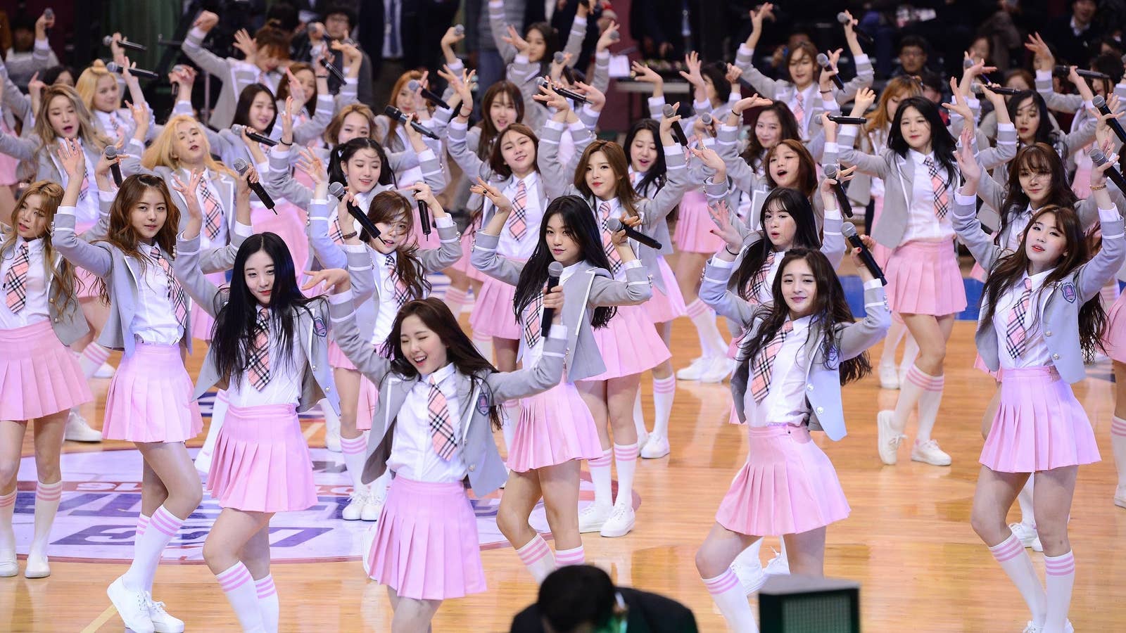 Dozens of K-pop girls wear skirts, ties, and blazers and pose for the camera while holding microphones