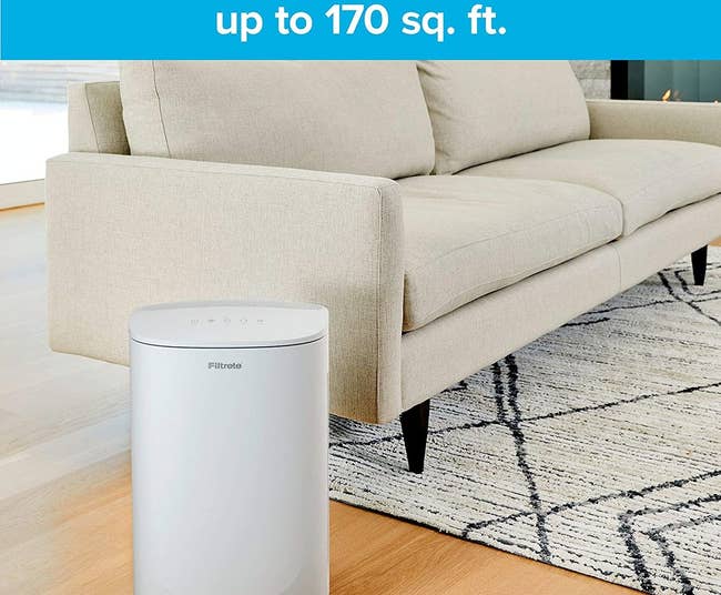 A white air purifier about the size of a of medium trash can next to a couch in a living room 