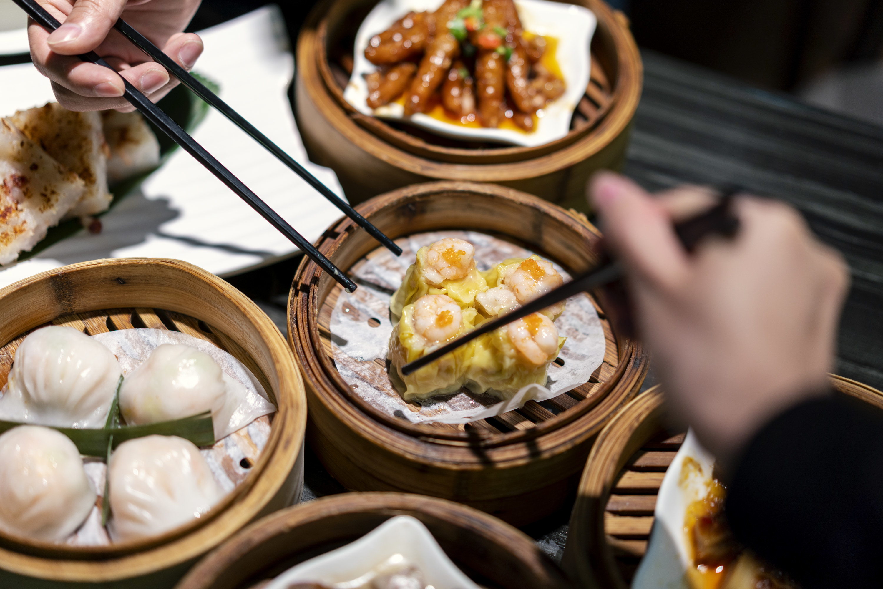 Dim sum sitting in multiple bamboo steamers with two pairs of hands picking up food with their chopsticks