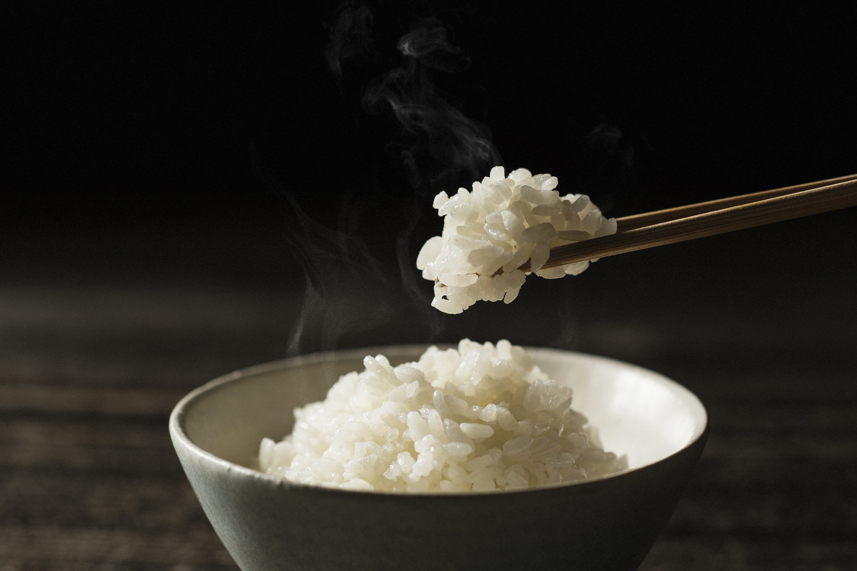 Steaming bowl of white rice with a pair of chopsticks holding a clump of rice