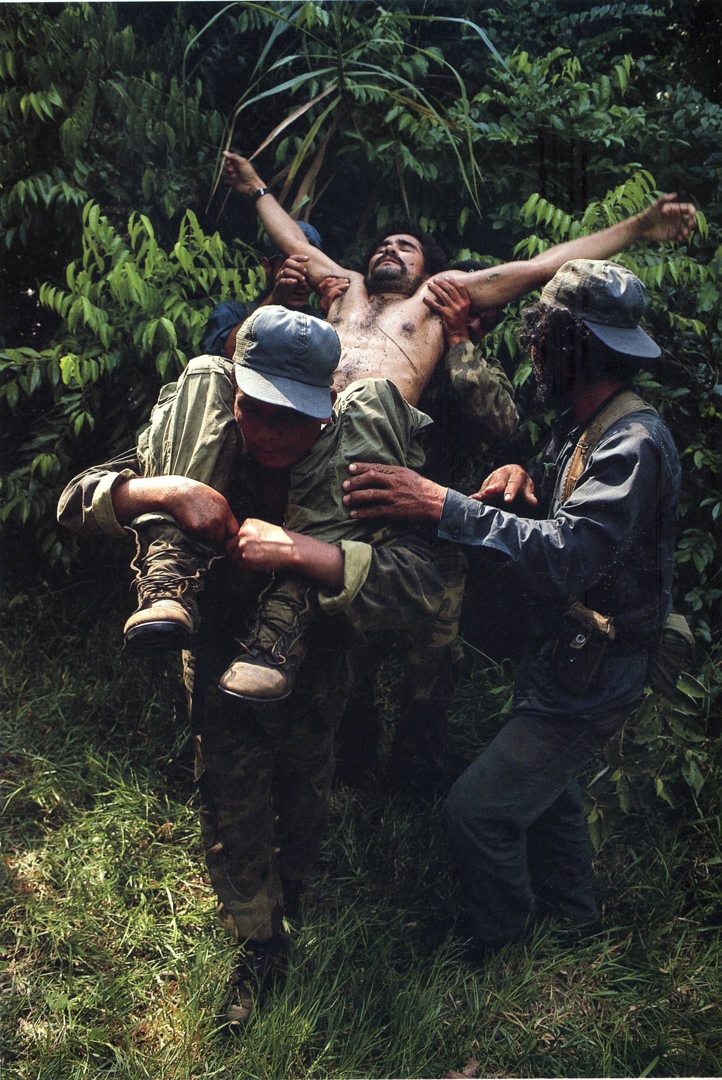 A dead or injured man is carried out of a jungle setting by soldiers with his arms extended, looking like Christ on the cross