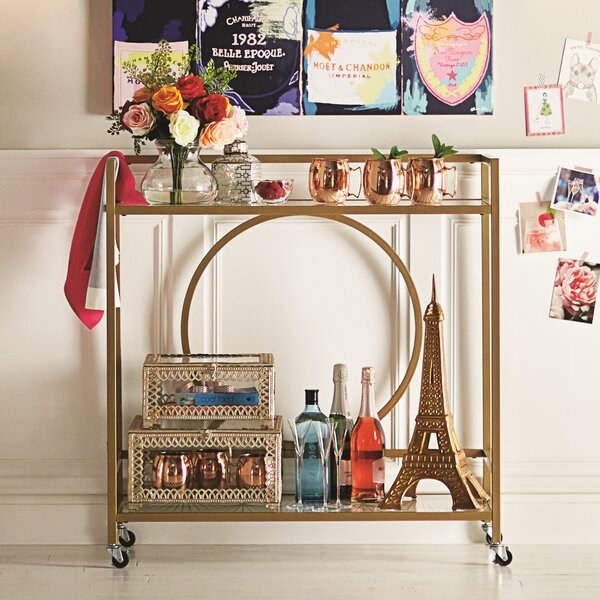 gold metal bar cart with wheels, with flowers and copper mugs on the top shelf and bottles and other decor on the bottom shelf
