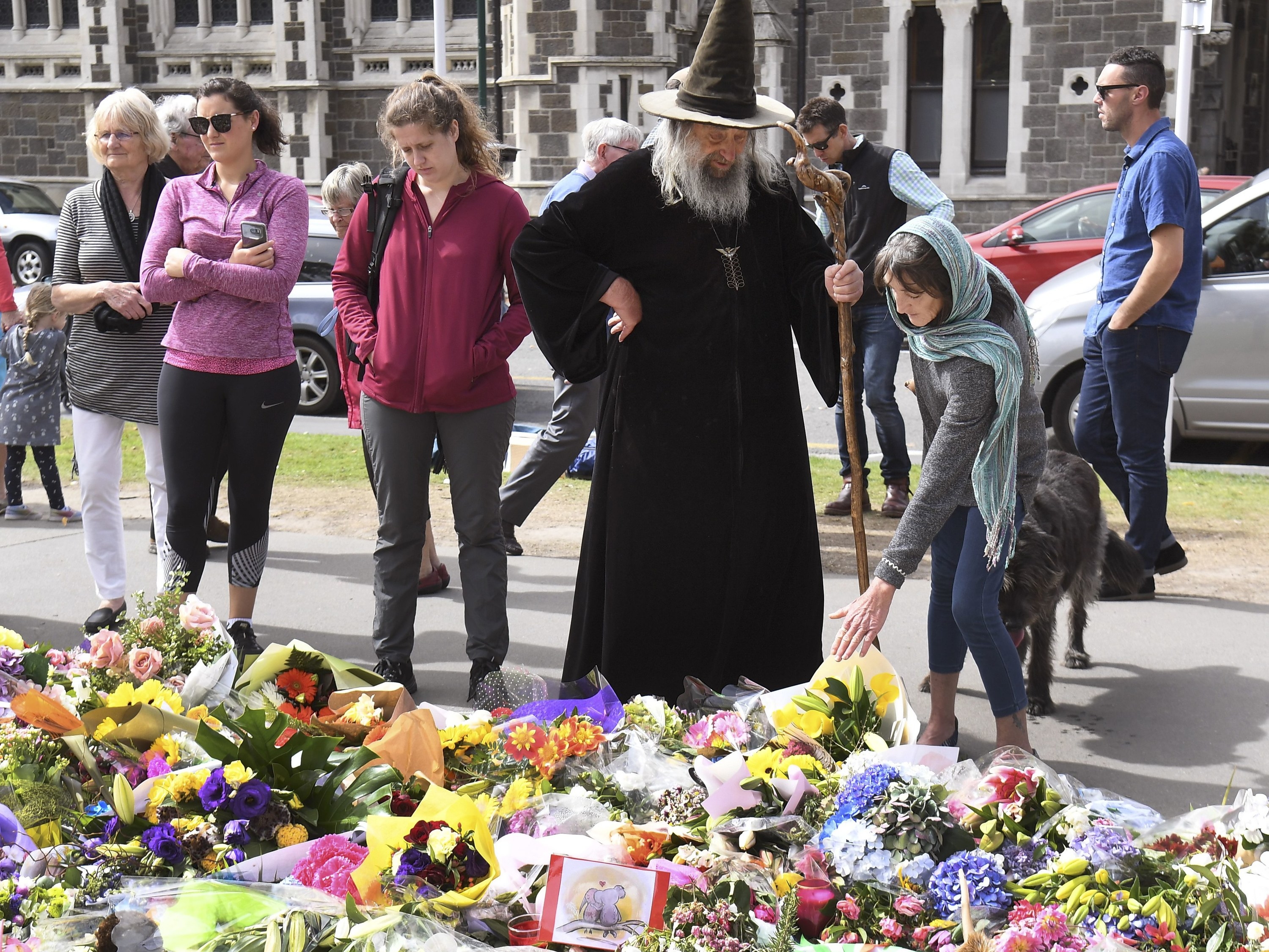Wizard wearing dark robes standing in front of a memorial blanketed with flowers. There are many people surrounding him