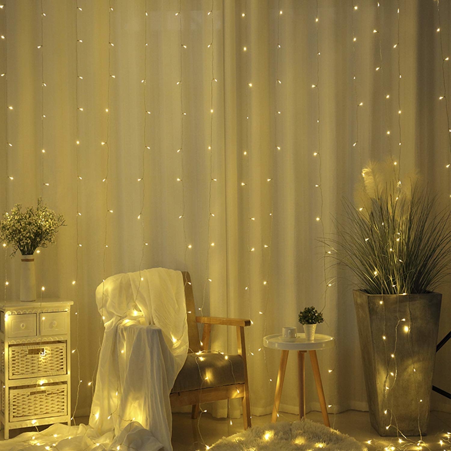 The light curtain layered with translucent white curtains. There&#x27;s a chair, plant, side table, and a cabinet in front of the curtains.