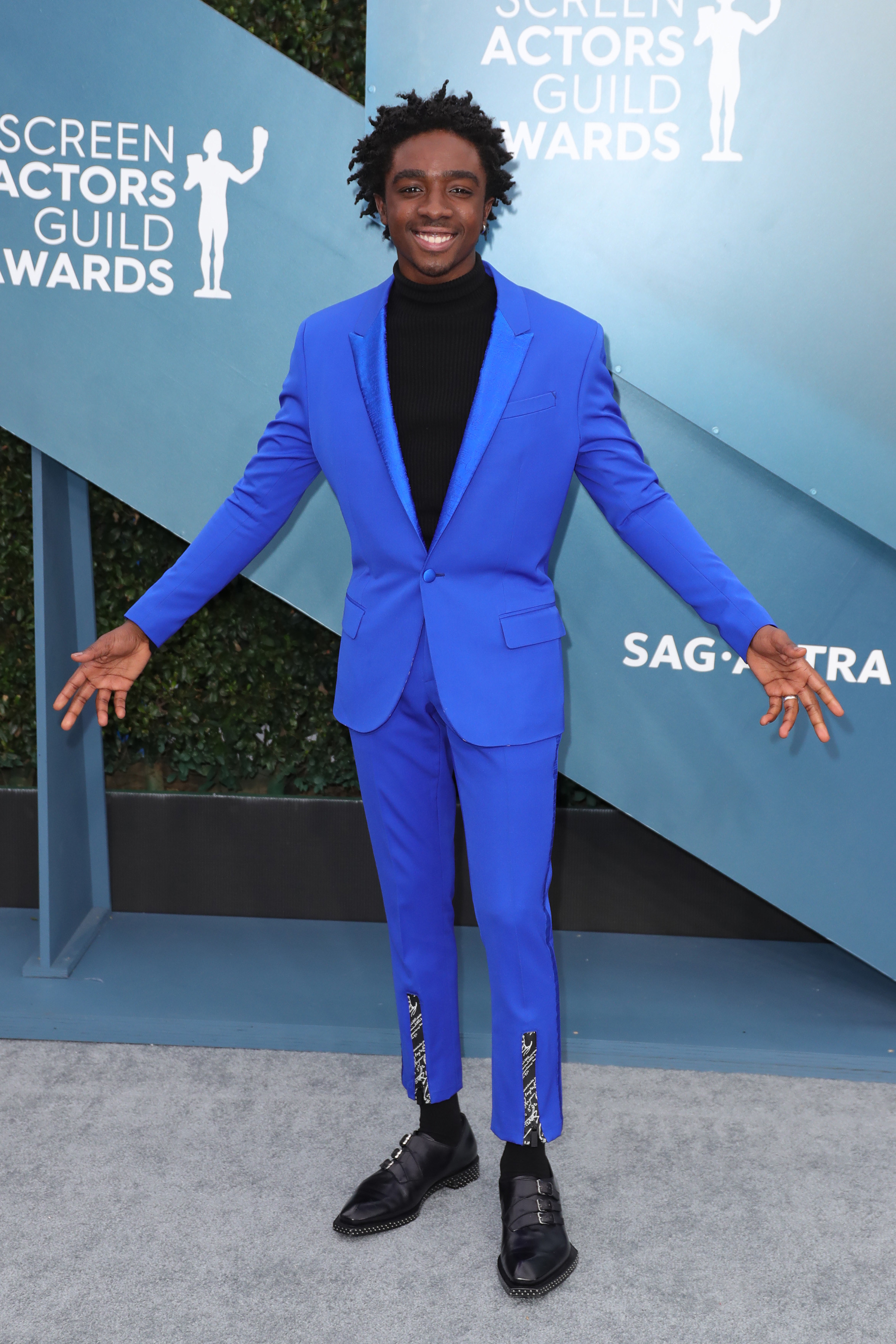 Caleb is holding his arms out on a red carpet to show off his monochromatic suit. 