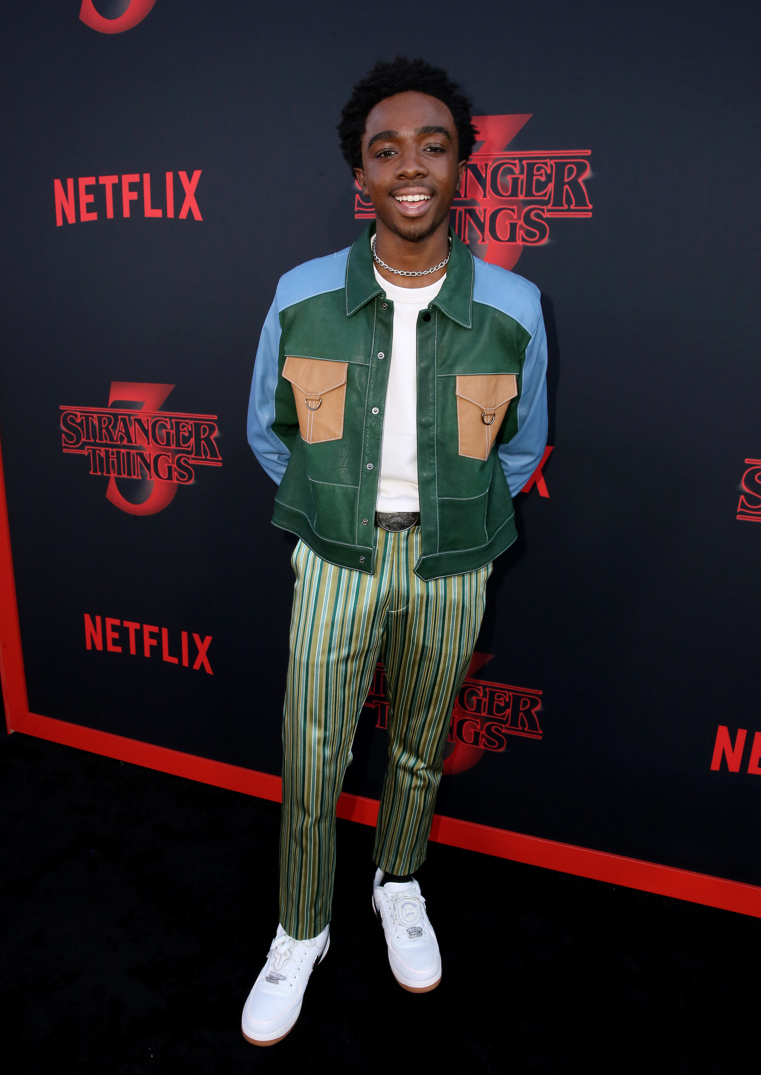 Caleb is smiling at a premiere wearing a multicolored jacket and printed pants. 