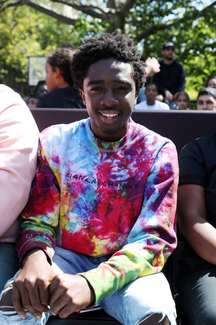 Caleb is smiling while wearing a multi-colored sweatshirt and distressed jeans. 