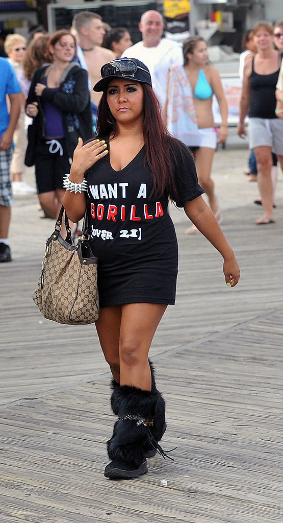 Snooki on a boardwalk wearing a graphic tee, tight skirt, furry boots, and a baseball cap