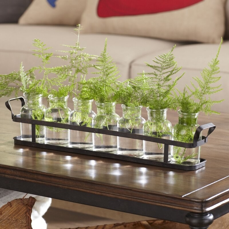 Metal tray with seven glass jars filled with greenery, sitting on a table