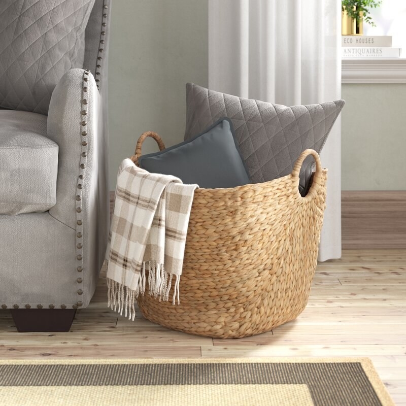 wicker basket with handles sitting on the floor filled with pillow and blankets