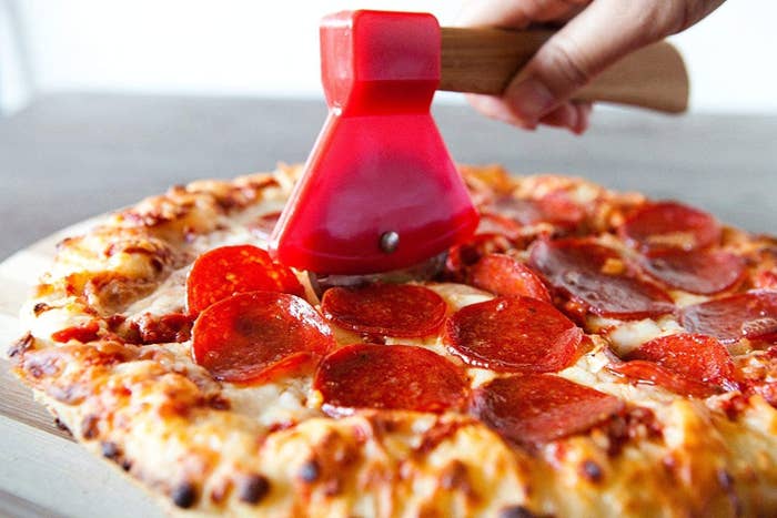 Hand using pizza cutter shaped like an axe to cut a pepperoni pizza