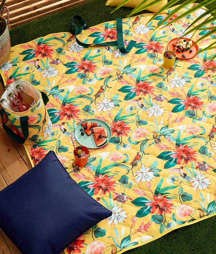 A floral outdoor blanket outside with fruits and beverages sitting on top of it
