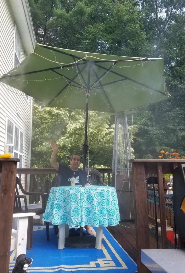 a reviewer sits on their patio under an umbrella with the orbit outdoor mist cooling system attached to it and spraying mist