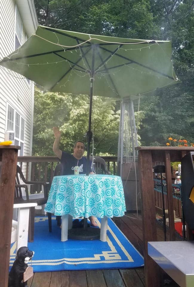 a reviewer sits on their patio under an umbrella with the orbit outdoor mist cooling system attached to it and spraying mist